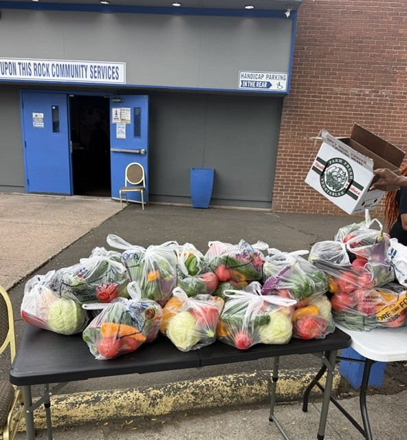 Tables of produce bags outside the Upon This Rock Community Services entrance as a volunteer unloads a farm-fresh box